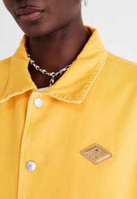 Gul denimjakke med krage, to knapper og et diamantformet lærmerke. Har synlige sømmer og en teksturert stoff.