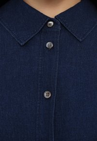 Gros plan sur le col d'une chemise en denim bleu foncé avec trois boutons gris visibles attachés le long de la patte de boutonnage.