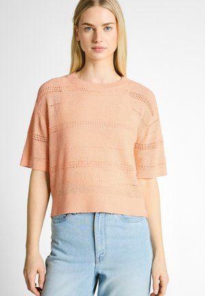 Jeune femme aux longs cheveux blonds portant un top en tricot pêche à manches courtes et un jean taille haute bleu clair, debout sur un fond blanc.