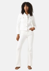 Chaqueta de denim blanca con dos bolsillos en el pecho, botones a presión y mangas largas, combinada con jeans blancos de campana y sandalias marrones con tiras.