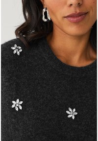 Donkergrijze trui met een gestructureerde afwerking, voorzien van zilver versierde bloemdetails. Stoere zilveren oorringen maken de look compleet.