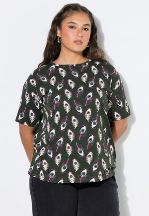 Mujer con el pelo largo que lleva una camisa verde de manga corta con un patrón de plumas de pavo real blancas y moradas, de pie con las manos detrás de la espalda.