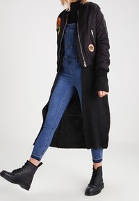 Svart bomberjacka med lappar, tillsammans med mörkblå overaller och svarta ankelboots. Lång svart cardigan med ribbad textur.