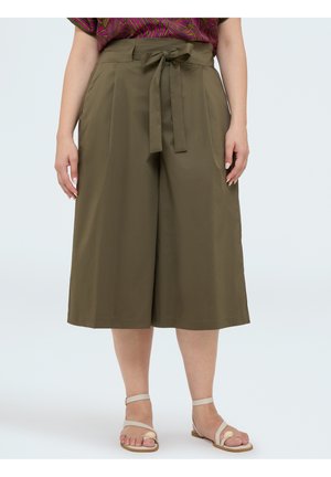 Femme portant un pantalon large court vert olive avec ceinture nouée à l'avant et des sandales beiges à brides, debout sur un fond uni.