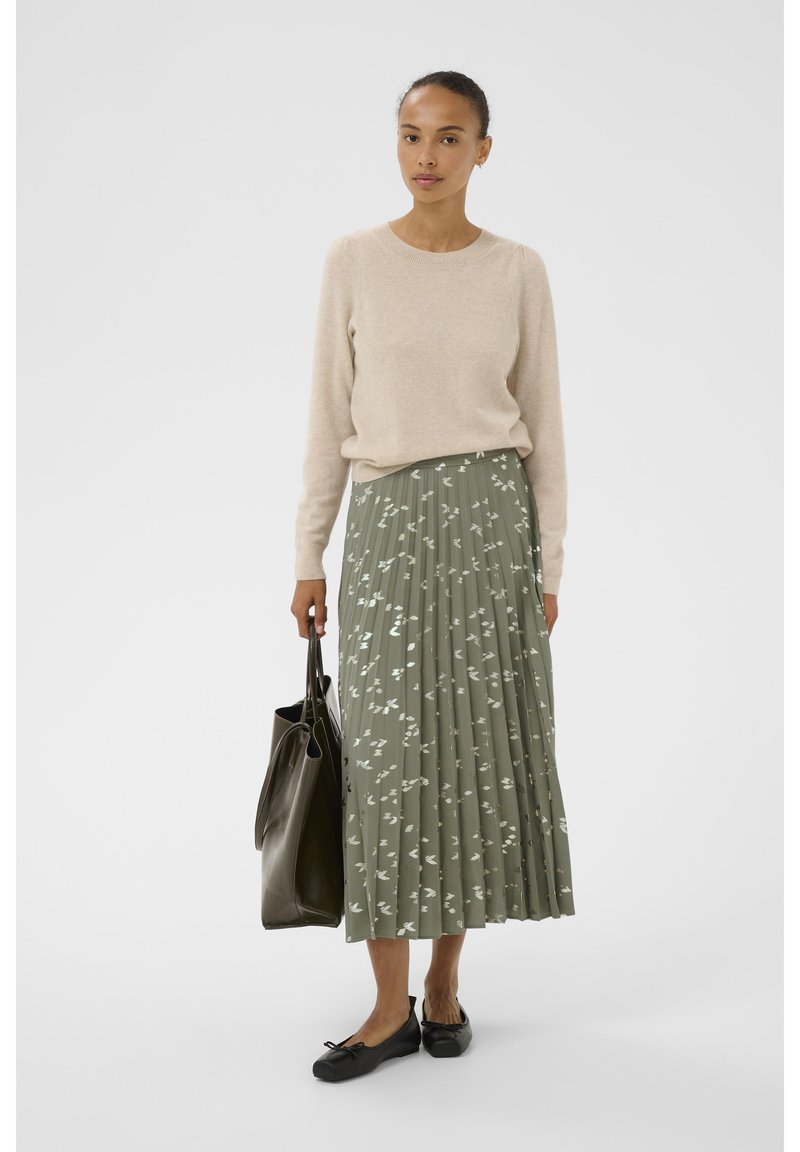 Maglione beige, gonna plissettata verde con motivo a foglie bianche, ballerine nere, in mano una borsa tote scura. Design semplice e strutturato.
