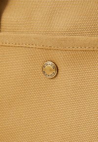 Gros plan sur un tissu texturé couleur tabac avec une couture horizontale et un bouton métallique circulaire gravé du mot "COACH".