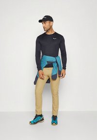 Schwarzes Sportshirt mit langen Ärmeln, beige Hose, türkisfarbene Jacke um die Taille gebunden, schwarze Mütze, blau-gelbe Turnschuhe.