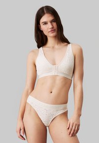 Ensemble bralette et culotte en beige, fabriqué en tissu texturé avec un motif subtil, comportant un fermoir à l'avant et une ceinture élastique avec logo.