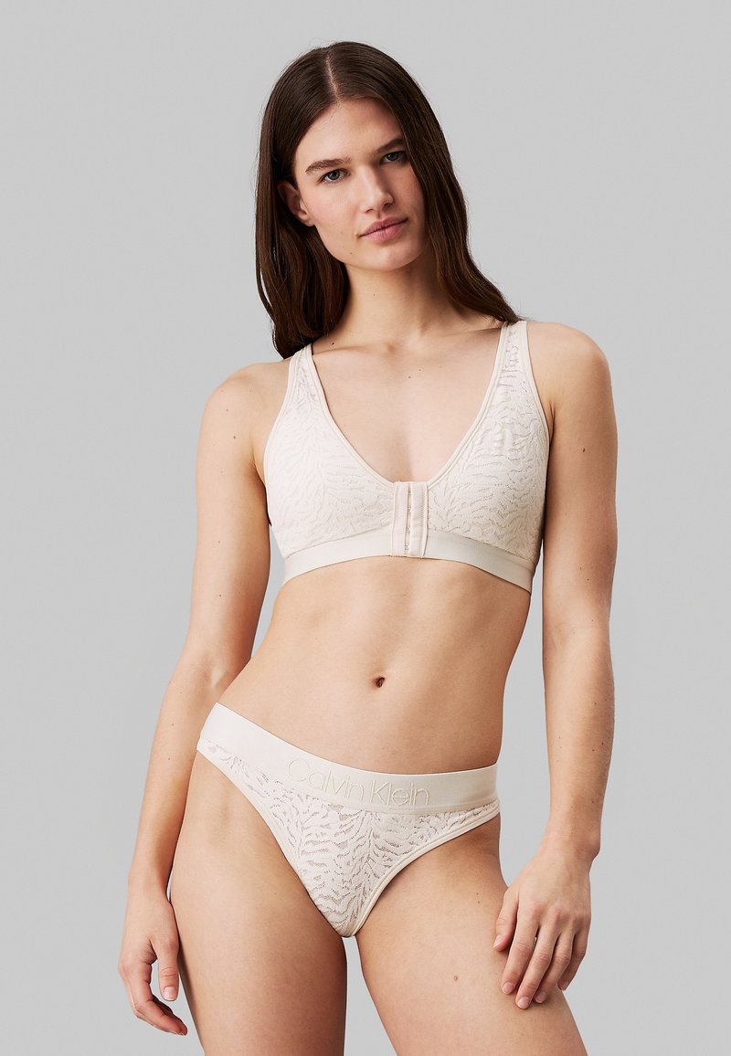 Ensemble bralette et culotte en beige, fabriqué en tissu texturé avec un motif subtil, comportant un fermoir à l'avant et une ceinture élastique avec logo.
