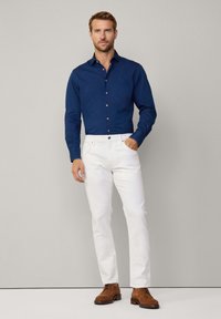 Marineblauw overhemd met knopen, witte jeans, bruine veterschoenen. Glad materiaal, slim fit en contrasterende knopen op het shirt.