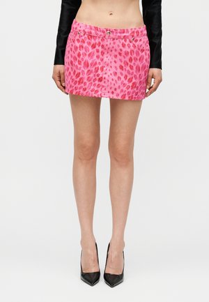 Frau trägt einen pinkfarbenen Minirock mit roten Leopardenflecken und schwarzen High Heels, steht vor einem schlichten weißen Hintergrund.