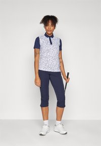 Golf-Polohemd mit einem weißen gemusterten Vorderteil und blauen Kurzärmeln. Kombiniert mit marineblauen Caprihosen und weißen Trainingsschuhen.