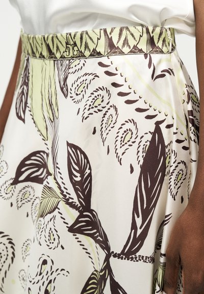 Κοντινό πλάνο μιας φούστας με εκρού φόντο που διαθέτει περίτεχνα σχέδια φύλλων και παϊλεϊν σε σκούρο καφέ και ανοιχτό πράσινο, συνδυασμένη με λευκό τοπ.