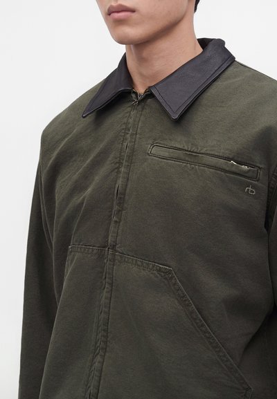 Homme portant une veste vert foncé avec un col noir et des poches visibles sur la poitrine et à l'avant, debout devant un fond clair uni.
