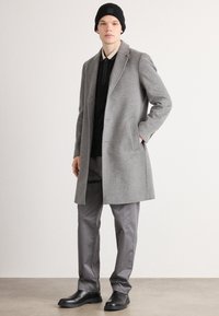 Cappotto di lana grigio con rever a scollatura, tasche laterali e una vestibilità su misura, indossato sopra un maglione nero e pantaloni grigi. Calzature nere completano il look.