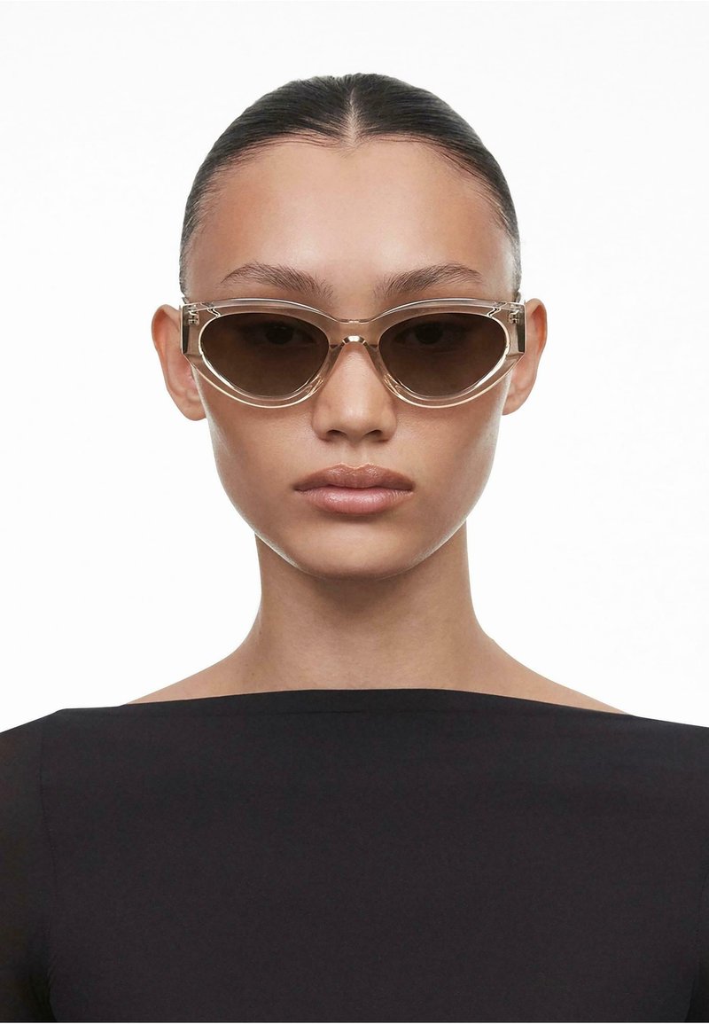Sunglasses med en klar, kattöga-ram och bruna tonade linser; har tjocka sidor med subtil branding. Modell bär en svart topp.