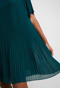 Vestido de color verde azulado con un corte plisado y una silueta fluida, que presenta una tela ligera y un efecto textural en capas, acentuado por mangas cortas.