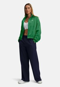 Groene sportjack met elastische manchetten, gecombineerd met een witte cropped top en marineblauwe wijde broek. Witte sneakers maken de outfit af.