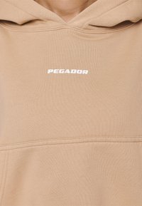 Beige hoodie med vit "PEGADOR" textlogotyp centrerad på bröstet och synliga sömnadsdetaljer.