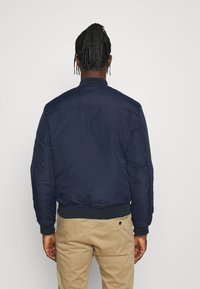 Giacca bomber blu navy con polsini e orlo a coste, caratterizzata da una superficie liscia e un design semplice. Collo regolabile, senza loghi visibili.