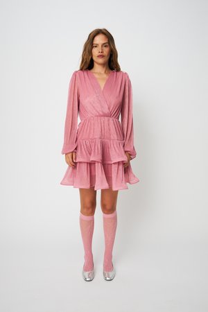 Γυναίκα που φοράει ένα ροζ μίνι φόρεμα με μακριά μανίκια και βαθμίδες, με ροζ καλσόν μέχρι το γόνατο και ασημένια παπούτσια, στέκεται μπροστά σε απλό φόντο.