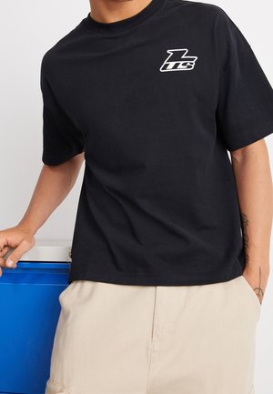 T-shirt en coton noir à manches courtes et col rond, avec un logo blanc sur la poitrine gauche. Porté avec un short beige.