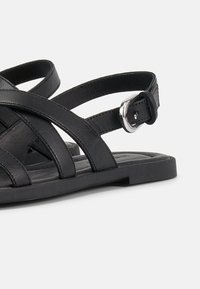 Μαύρο επίπεδο σανδάλι με πολλαπλές λουρίδες και ασημένια αγκράφα σε λουρί τύπου slingback, εμφανιζόμενο σε λευκό φόντο.