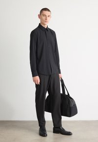 Sort knappskjorte med lange ermer, kombinert med sorte bukser og lærskot; holder en svart duffelbowe. Enkel, stilren design.