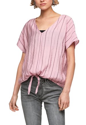 Woman wearing a pink short-sleeve V-neck blouse with vertical stripes and a front tie, paired with faded gray jeans and silver earrings.