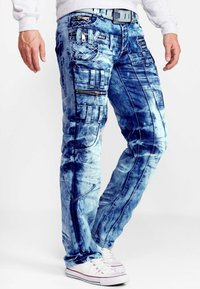 Jeans in denim blu con un design sbiadito e strappato, caratterizzati da molteplici tasche, cerniere e dettagli di cucitura. Abbinati a sneaker bianche.