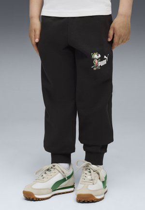 Pantalones de chándal negros con puños elásticos, con un logotipo bordado colorido y un personaje de dibujos animados; usados con zapatillas blancas y verdes.