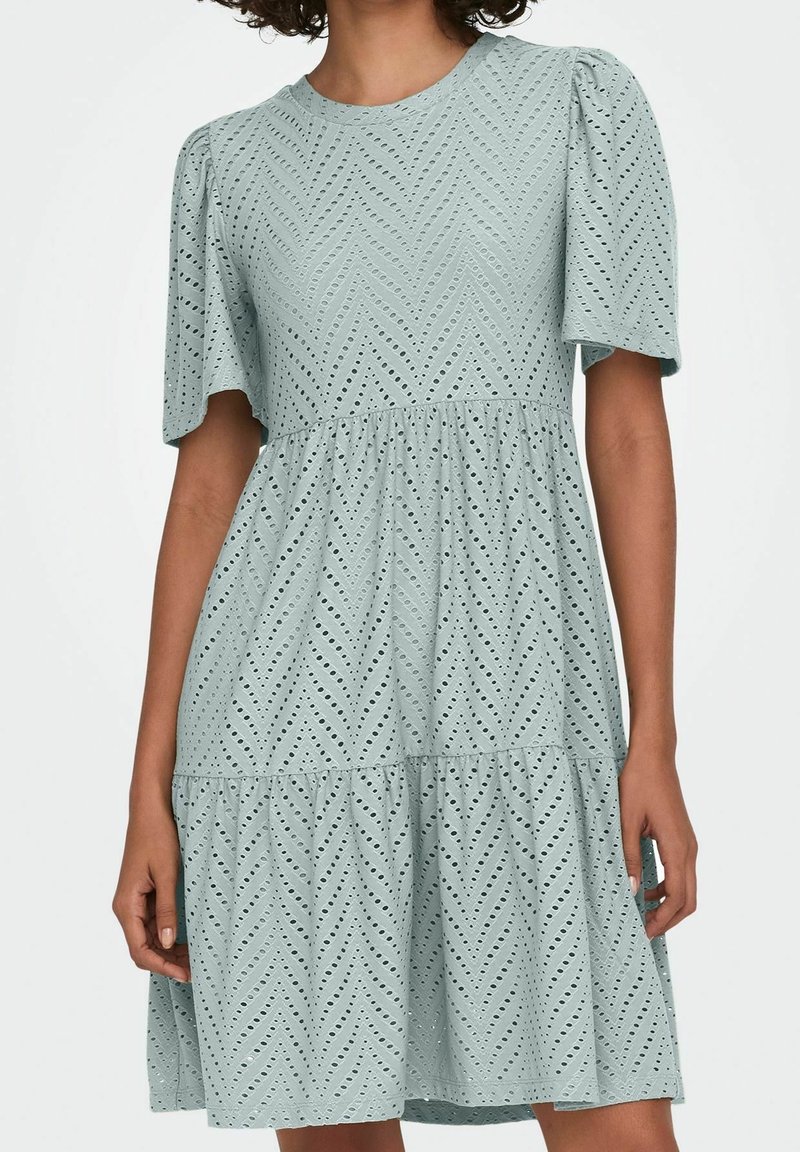 Femme portant une robe bleu clair à œillets avec manches courtes bouffantes et jupe à volants.