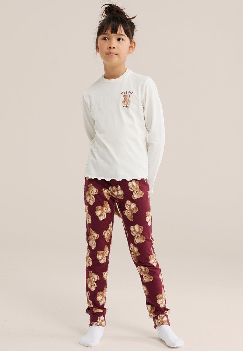 Enfant debout portant un haut blanc à manches longues avec un ours en peluche et du texte, ainsi qu'un pantalon bordeaux couvert d'imprimés d'ours en peluche et des chaussettes blanches.