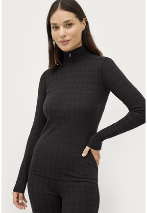 Kvinde iført en tætsiddende sort langærmet top med høj lynlås-krave og subtilt tekstureret mønster, der poserer med den ene hånd på hoften.