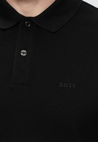 Fekete galléros póló két gombbal begombolva, és finoman hímzett "BOSS" logóval a bal mellrészen.