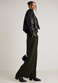 Μαύρο cropped δερμάτινο τζάκετ συνδυασμένο με χαλαρές, ελιάς πράσινες παντελόνες. Μοντέλο που φοράει μαύρες μποτάκια με τακούνι και κρατάει μια μικρή τσάντα, σε ουδέτερο φόντο.