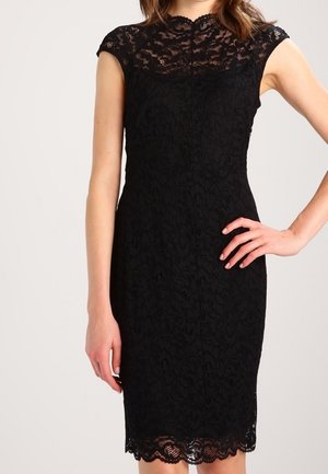 Femme portant une robe noire en dentelle ajustée avec des manches courtes et un ourlet festonné, posant avec une main sur la hanche devant un fond uni.