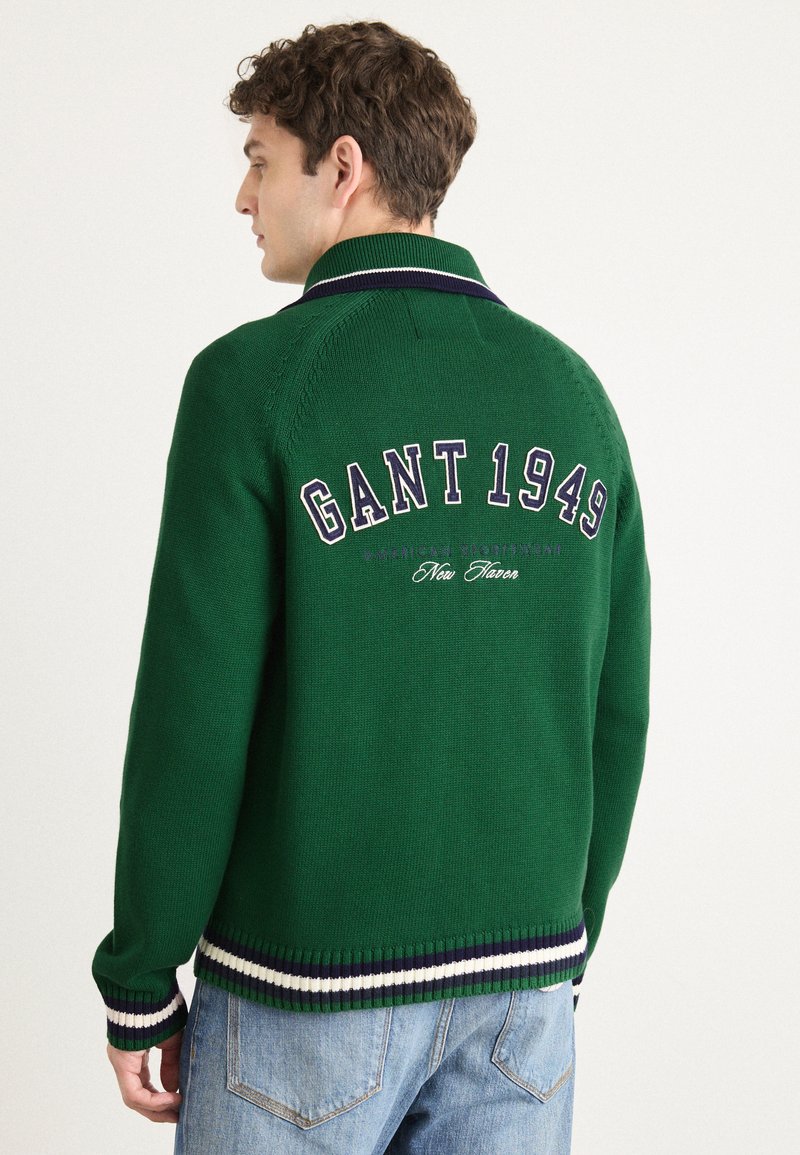 Groene gebreide trui met een kraagontwerp, voorzien van witte en marineblauwe gestreepte accenten aan de onderkant en mouwen, en "GANT 1949" geborduurd op de achterkant.