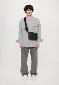 Crna pravokutna crossbody torba s teksturiranim finišom, podesivim remenom, postavljena na modelu koji nosi prugastu košulju na dugmadi i sive hlače.