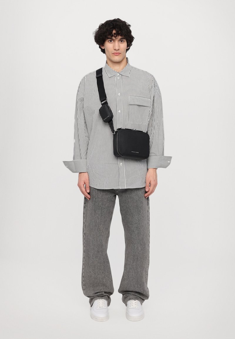 Crna pravokutna crossbody torba s teksturiranim finišom, podesivim remenom, postavljena na modelu koji nosi prugastu košulju na dugmadi i sive hlače.