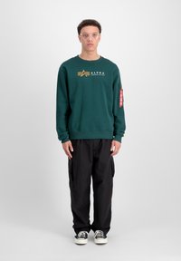 Tamnozeleni dukserica s zlatnim logotipom i crvenom oznakom na rukavu, u kombinaciji s crnim cargo hlačama i crno-bijelim tenisicama.