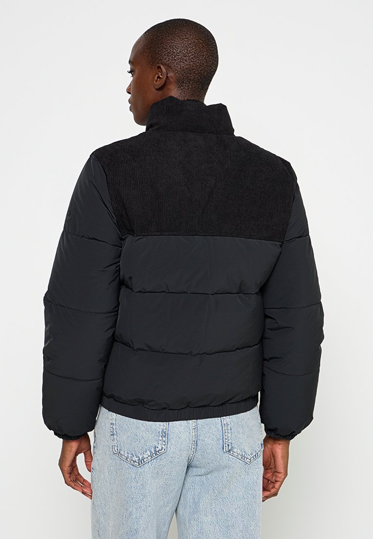 Schwarze Pufferjacke mit einem strukturierten oberen Rückteil aus schwarzem Cordstoff, ausgestattet mit horizontaler Steppung und einem hohen Kragen. Ärmelsäume aus Baumwolle.