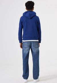 Sudadera azul con capucha y bolsillo canguro, combinada con jeans rectos de un azul claro y zapatillas blancas. Diseño simple y casual.