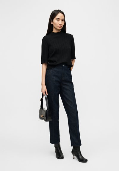 Μαύρο ριμπ πλ εκτό τοπ με κοντά μανίκια, σε συνδυασμό με σκουρόχρωμο τζιν παντελόνι με ίσια γραμμή. Το μοντέλο κρατά μια μικρή μαύρη τσάντα χειρός και φοράει μαύρα μποτάκια αστραγάλου.