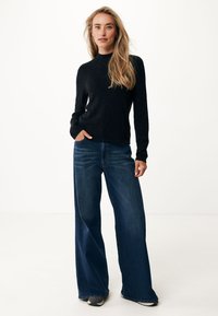Černý pletený svetr s dlouhými rukávy, v kombinaci s volnými modrými džíny. Texturovaná látka a uvolněný střih. Detail obuvi není uveden.