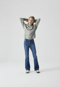 Pull en maille gris, à coupe ample et col large, associé à un jean bleu évasé et des baskets blanches. Tenue simple et décontractée sur fond neutre.