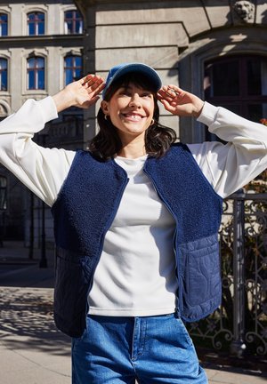 Mladá žena sa usmieva vonku a upravuje si modrú šiltovku, má na sebe bielu košeľu s dlhým rukávom, tmavomodrú vestu a modré džínsy v mestskom prostredí.