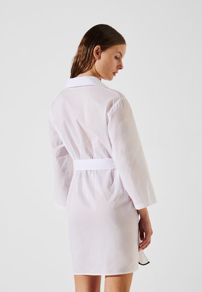 Robe en coton rose clair avec un col, des manches trois-quarts et une ceinture à nouer à la taille. Présente une texture lisse et une longueur mi-cuisse.