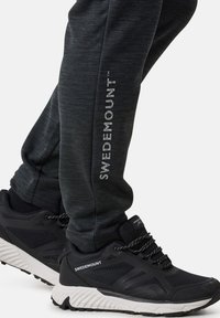 Schwarze Sporthosen mit melierter Textur, ausgestattet mit einem vertikalen "SWEDEMOUNT"-Logo. Kombiniert mit schwarzen Sneakern mit weißen Sohlen.
