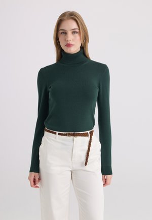Jeune femme aux cheveux blonds portant un pull à col roulé vert foncé, un pantalon blanc et une ceinture tressée marron sur un fond uni.